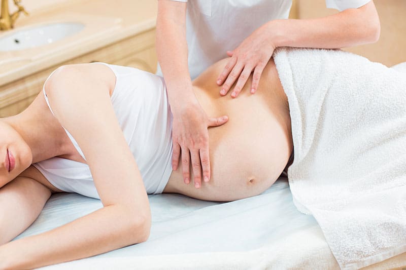 Close to the female hands of a massage therapist makes a light massage to the belly of a pregnant girl in a cosmetology room