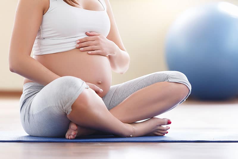Portrait of a beautiful young pregnant woman holding her stomach while sitting near an exercise ball in the gym. Working out and fitness, pregnancy concept